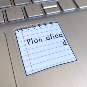 A sticker which reads plan ahead, on a lined paper notebook, stuck to a laptop
